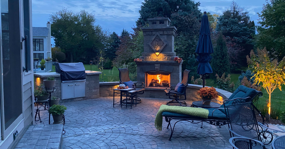 Stunning night view of a paver patio featuring an orange glow from a custom outdoor fireplace.