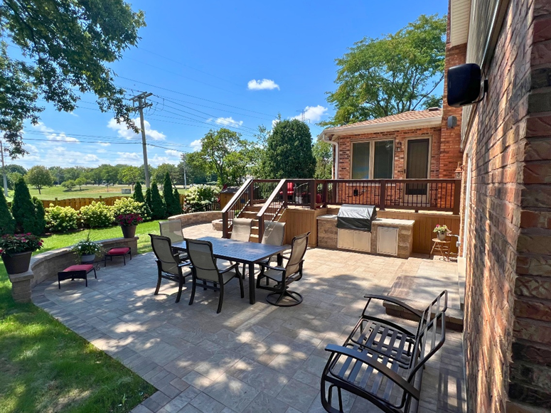 Custom paver patio with dining area and adjacent raised deck featuring a built-in outdoor kitchen.