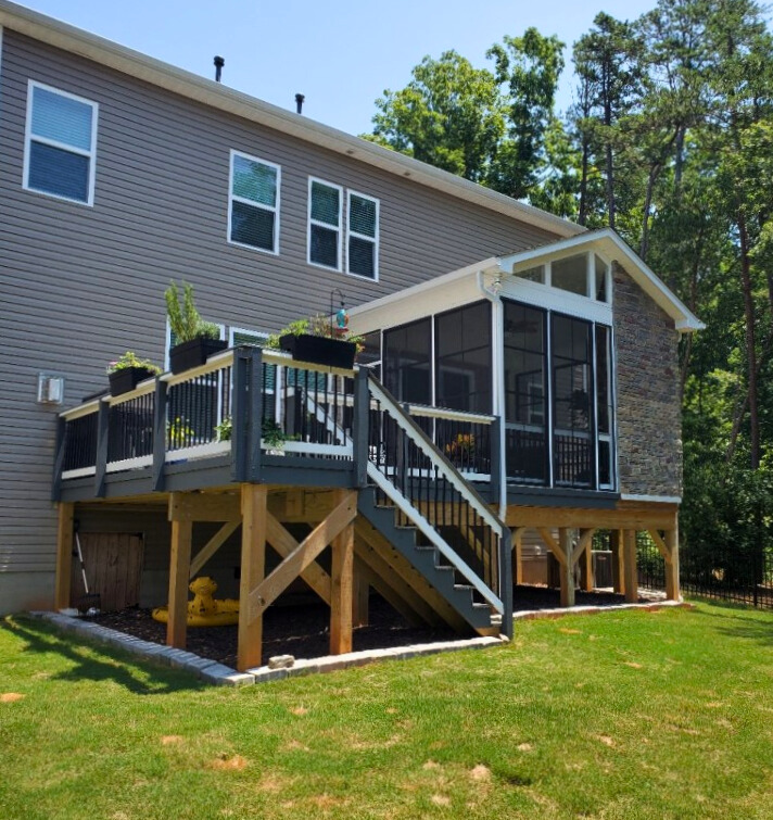 A two-story gray house with a new elevated wooden deck painted gray, leading down to a screened porch with a stone accent wall and stairs.