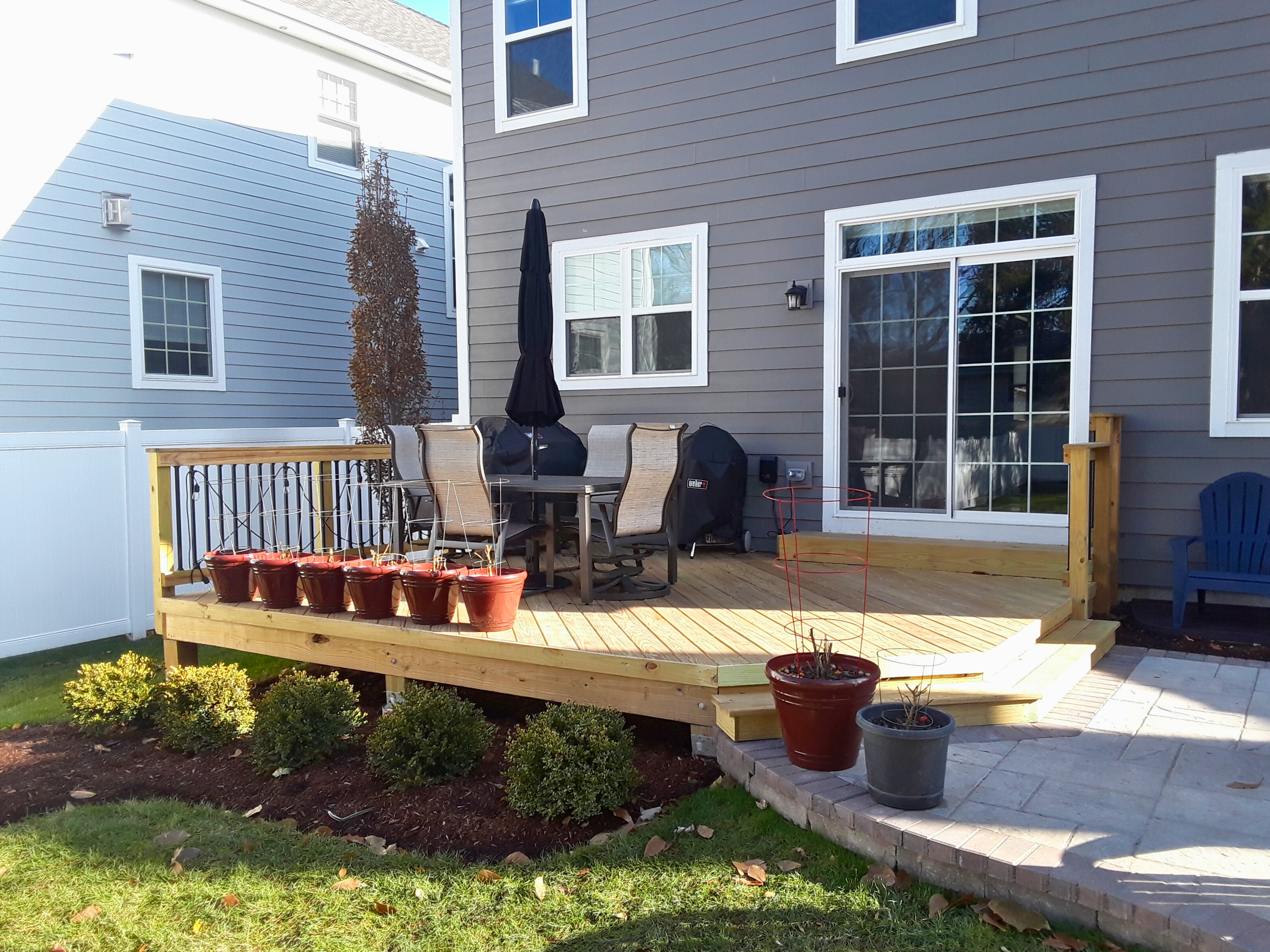 Ground-level wooden deck with steps leading to a paver patio next to a gray house in Downers Grove, IL. This combination outdoor living space, built by Archadeck of Chicagoland, features a seating area with an umbrella.