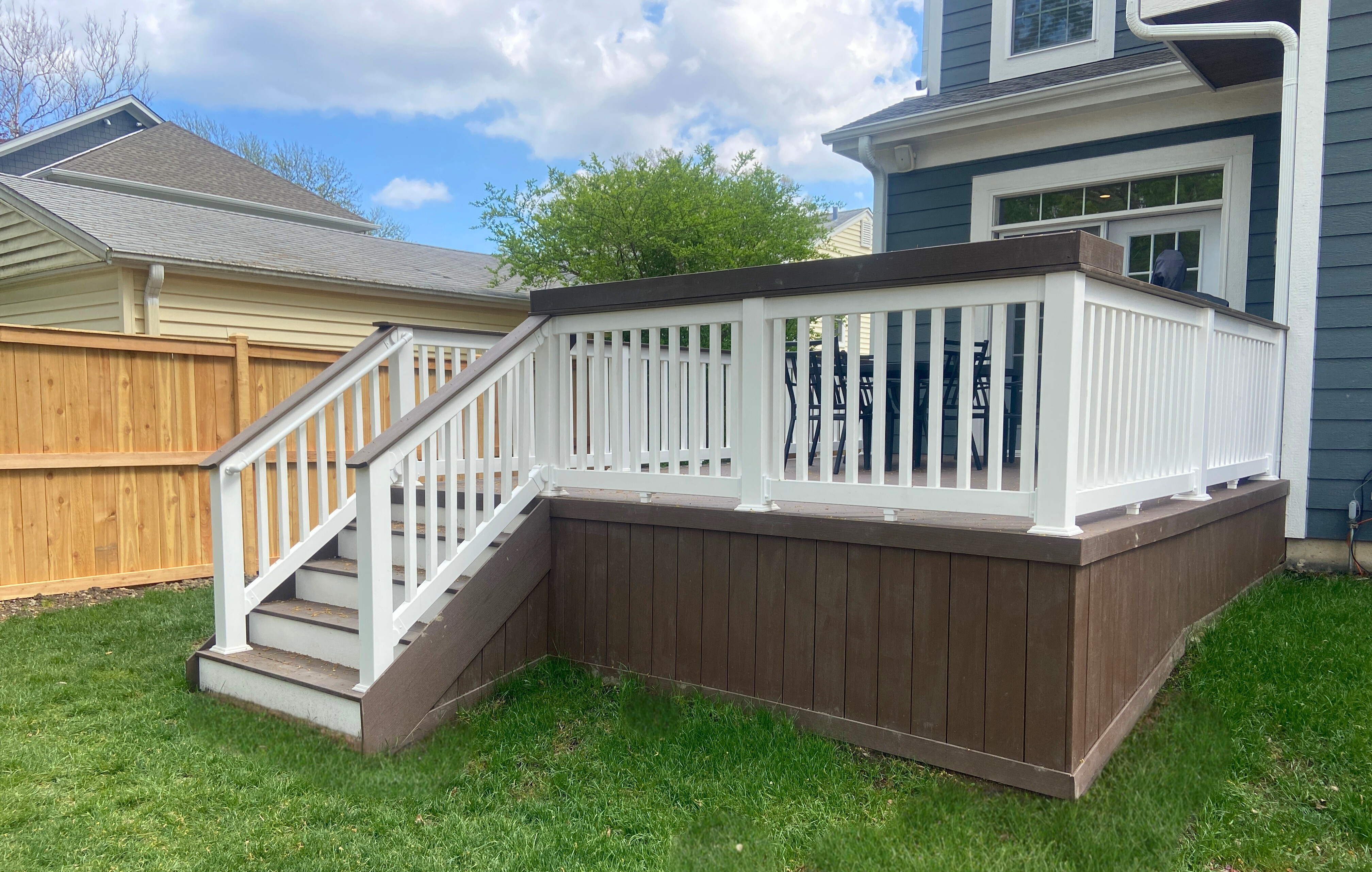 A modern composite deck with dark brown planks and elegant white railings, featuring steps leading down to a green lawn, attached to a dark blue-sided home in Deerfield, IL.